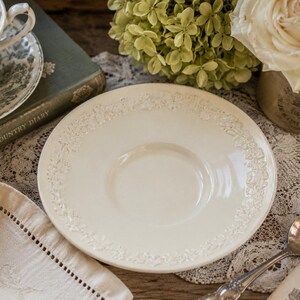 May include: Cream-colored saucer with a raised floral pattern around the rim. The saucer is part of a vintage tea set, with a matching teacup visible in the background. The Country Diary book is also in the background.