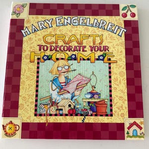 Może przedstawiać: Kwadratowa książka w twardej oprawie zatytułowana Mary Engelbreit Crafts to Decorate Your Home. Okładka ma żółte kwiatowe tło z bordową szachownicą oraz ilustrację osoby zajmującej się rękodziełem przy użyciu różnych narzędzi.