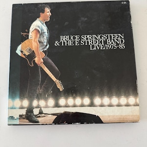 May include: A square vinyl record box set titled Bruce Springsteen & The E Street Band Live/1975-85, featuring a black and white photograph of a musician playing an electric guitar on stage with a row of bright stage lights at the bottom.