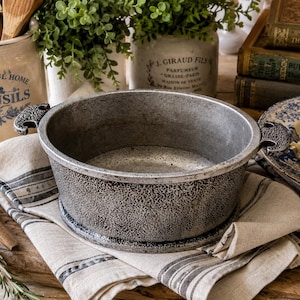 May include: A textured, silver-colored cast iron pot with handles, resting on a striped linen cloth. The pot is surrounded by kitchen utensils, greenery, and vintage books, suggesting a rustic, farmhouse kitchen theme.