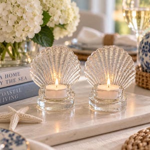 May include: Two clear glass tea light candle holders shaped like scalloped seashells. Each holder features a textured, fan-shaped back panel and a circular base that supports a small, round white tea light candle with a glowing flame.