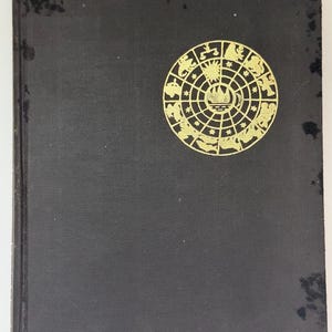 May include: A vintage hardcover book with a black cover and a gold circular emblem featuring zodiac symbols. The book's cover shows signs of wear, adding to its antique charm. The emblem is centered on the cover.
