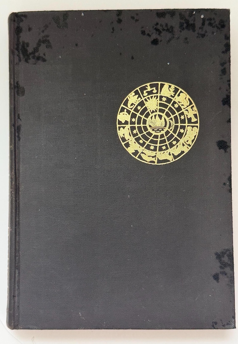 May include: A vintage hardcover book with a black cover and a gold circular emblem featuring zodiac symbols. The book's cover shows signs of wear, adding to its antique charm. The emblem is centered on the cover.