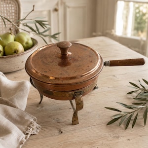 May include: A round copper cooking pot with a matching lid, a wooden handle, and three curved metal legs. The pot features a weathered metallic finish with visible signs of use, a central knob on the lid, and a sturdy base structure.