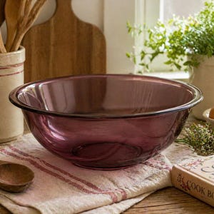 Puede incluir: Un gran cuenco de vidrio morado transparente se encuentra sobre una toalla de lino a rayas. Una cuchara de madera descansa cerca. Otros artículos de cocina incluyen un libro de cocina, utensilios de madera y un recipiente de cerámica.