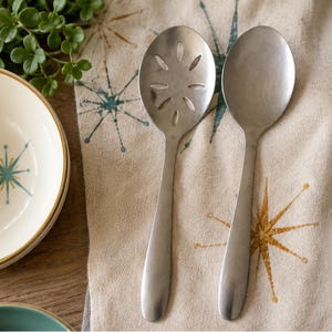 May include: Two silver-toned metal serving spoons, one featuring a solid oval bowl and the other featuring an oval bowl with a decorative floral-shaped cutout pattern. Both spoons have smooth, rounded handles and a matte finish.