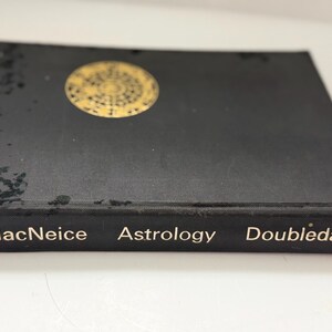 May include: A black book titled "Astrology" by MacNeice, published by Doubleday. The book has a gold circular design on the cover. The book is on a white surface.