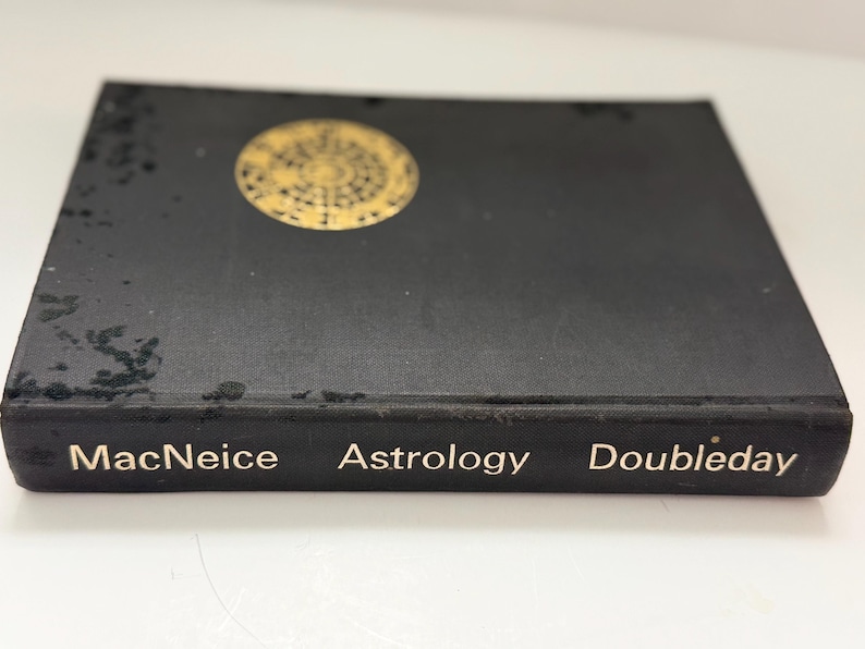May include: A black book titled "Astrology" by MacNeice, published by Doubleday. The book has a gold circular design on the cover. The book is on a white surface.