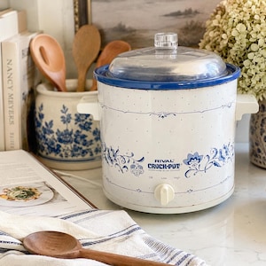 May include: A vintage Rival Crock-Pot slow cooker in white with blue floral accents and a clear lid. The appliance has a blue rim and a control knob. A wooden spoon and cookbooks are in the background.