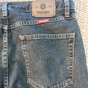 K&ouml;nnte beinhalten: Nahaufnahme der R&uuml;ckseite einer blauen Wrangler-Jeans. Die Jeans hat eine Ges&auml;&szlig;tasche und einen braunen Lederaufn&auml;her mit dem Wrangler-Logo. Der Aufn&auml;her lautet "Premium Quality" und "Authentic Jeans."