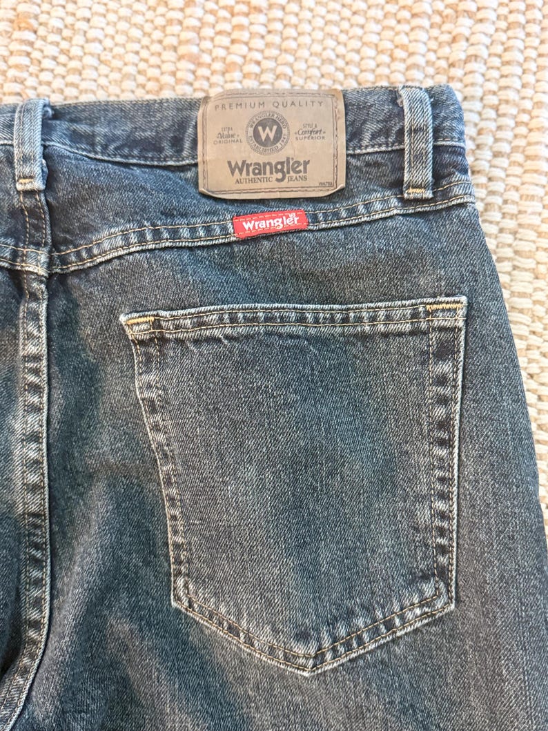 K&ouml;nnte beinhalten: Nahaufnahme der R&uuml;ckseite einer blauen Wrangler-Jeans. Die Jeans hat eine Ges&auml;&szlig;tasche und einen braunen Lederaufn&auml;her mit dem Wrangler-Logo. Der Aufn&auml;her lautet "Premium Quality" und "Authentic Jeans."