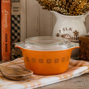 May include: A round, bright orange glass casserole dish with a clear glass lid. The exterior of the dish features a repeating horizontal band of brown, star-shaped snowflake patterns. The dish has two integrated handles on opposite sides.