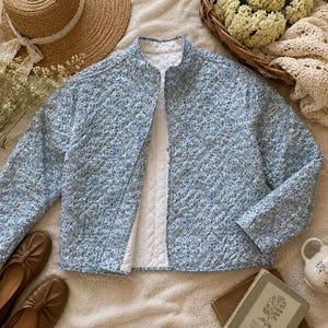 May include: A light blue quilted jacket with a floral pattern. The jacket has a round collar and a white lining. Brown leather flats are visible in the foreground, along with a straw hat and a small ceramic mug.