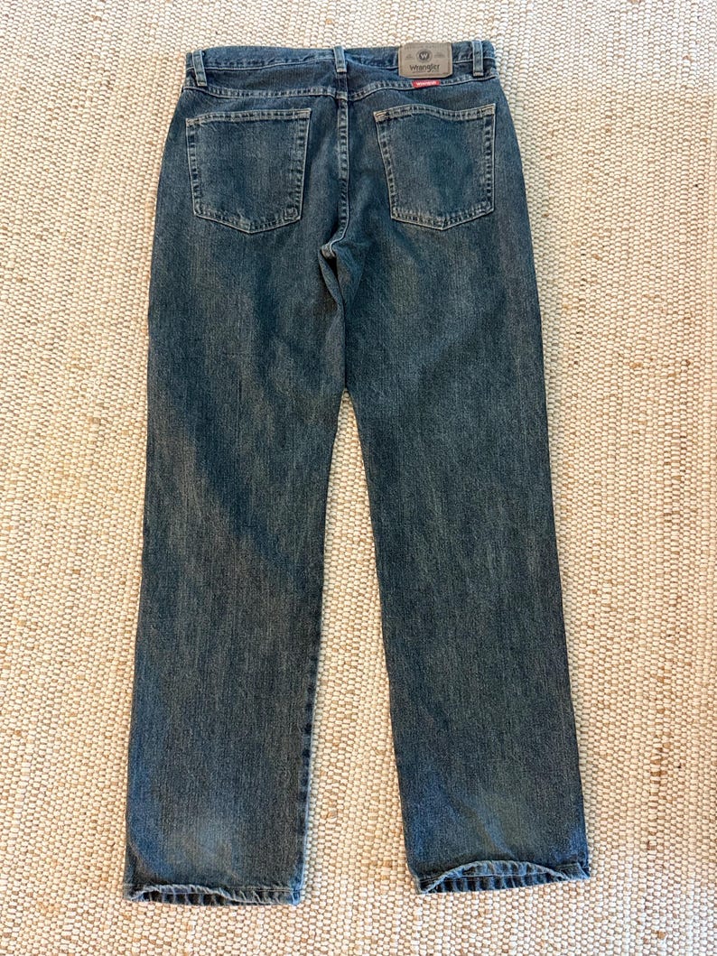 K&ouml;nnte beinhalten: R&uuml;ckansicht einer blauen Jeans mit zwei Ges&auml;&szlig;taschen und einem Wrangler-Etikett. Die Jeans hat ein klassisches F&uuml;nf-Taschen-Design und eine gerade Beinpassform. Der Denim hat einen verblichenen, abgenutzten Look.