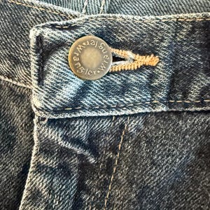 K&ouml;nnte beinhalten: Nahaufnahme einer blauen Jeans. Der Metallknopf ist mit dem Wort "Wrangler" gepr&auml;gt. Die Jeans hat sichtbare N&auml;hte und einen Knopfverschluss.