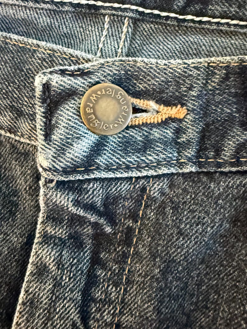 K&ouml;nnte beinhalten: Nahaufnahme einer blauen Jeans. Der Metallknopf ist mit dem Wort "Wrangler" gepr&auml;gt. Die Jeans hat sichtbare N&auml;hte und einen Knopfverschluss.