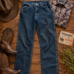 K&ouml;nnte beinhalten: Draufsicht auf Western-Kleidung, darunter blaue Jeans, ein kariertes Button-Down-Hemd, braune Cowboy-Stiefel und ein brauner Cowboyhut. Ein Vintage-Wrangler-Schild ist ebenfalls vorhanden.