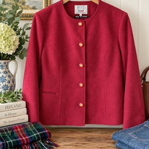 May include: A solid deep red jacket with a round neckline and long sleeves. The front features five circular buttons with gold centers and red rims. A white label at the inner neck reads Avoca Collection Made in Ireland with a small graphic icon.