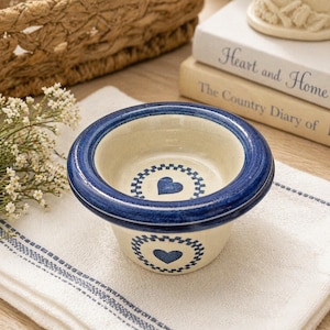 Op de afbeelding: Een kleine, crèmekleurige keramische kom met een blauwe rand en hartvormgeving. De kom heeft een blauw hart in het midden en een decoratieve rand. De kom staat op een wit en blauw gestreepte doek.