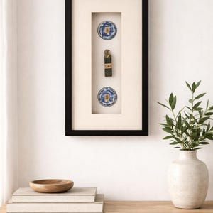 May include: Framed artwork featuring a black rectangular frame with a cream-colored mat. The frame holds two small blue and white decorative plates and a dark, cylindrical object. A wooden bowl and books sit on a wooden surface.