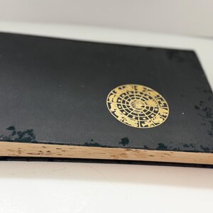 May include: A black book with a gold circular design on the cover. The design features an intricate pattern of shapes and symbols. The book's pages have a yellowed appearance, suggesting age. The book is slightly angled, revealing its thickness.