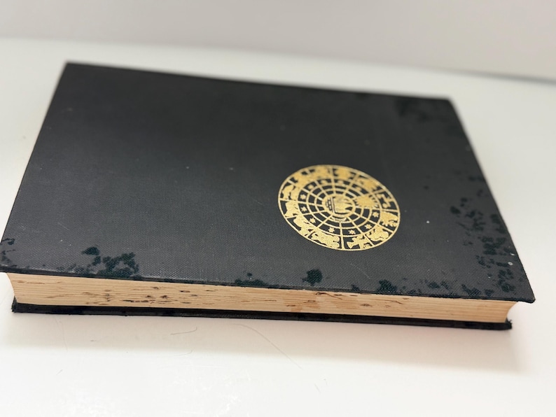 May include: A black book with a gold circular design on the cover. The design features an intricate pattern of shapes and symbols. The book's pages have a yellowed appearance, suggesting age. The book is slightly angled, revealing its thickness.