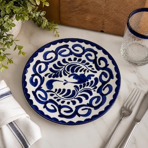 May include: A white ceramic plate with a blue floral design and a scalloped edge. The plate is part of a table setting that includes a clear glass with a blue rim, cutlery, and a folded napkin.