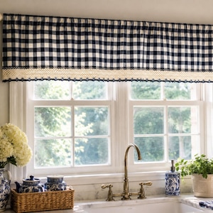 May include: A navy blue and white gingham valance with a cream-colored lace trim hangs above a window. The window is framed by white trim and overlooks a lush green outdoor scene. The kitchen decor includes a floral vase and a wicker basket.