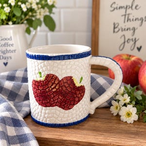 May include: A white ceramic mug featuring a textured mosaic-style surface with a blue rim and a central design of two red apples. The mug has a curved handle and a repeating grid pattern embossed across the exterior body of the vessel.
