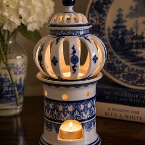 May include: A white ceramic wax warmer featuring a blue floral and geometric pattern. The cylindrical base has a cutout for a tea light candle, while the removable top is a dome-shaped lid with vertical cutouts, allowing light to shine through.