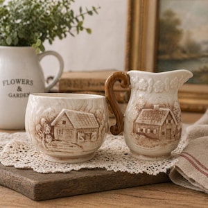 May include: Cream-colored ceramic cup and creamer set. Each piece features a raised relief design of a house and trees. The creamer has a wooden-look handle. The cup and creamer sit on a wooden board with a lace doily.