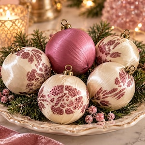 Könnte beinhalten: Ein Set aus fünf kugelförmigen Weihnachtskugeln mit glatter, satinartiger Oberfläche. Vier Kugeln sind cremefarben mit rosafarbenen Glitzer-Blumenmustern, während eine zentrale Kugel eine einfarbige, texturierte mauve-rosa Farbe aufweist.
