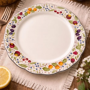 May include: White ceramic plate with a scalloped edge, decorated with a colorful fruit and berry design. The border features illustrations of peaches, plums, raspberries, and blueberries with green leaves. The plate is set on a beige linen napkin.