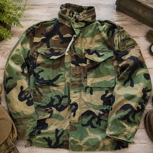 May include: A camouflage field jacket featuring a high collar, two chest pockets with buttoned flaps, and a shoulder patch with the text 1st Armored Division Old Ironsides. The jacket has a woodland pattern in shades of green, brown, and black.