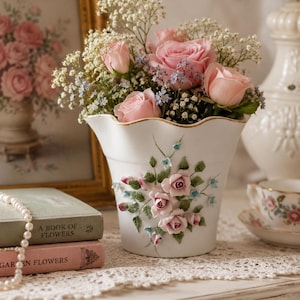 May include: A white ceramic vase with a scalloped gold-trimmed rim, featuring raised three-dimensional pink rose and green leaf decorations on the front, filled with a bouquet of fresh pink roses, white baby's breath, and small blue flowers.