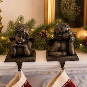 May include: A pair of bronze-toned metal stocking holders featuring cherub figures with wings. Each cherub rests on a rectangular base with a hook extending downward to support a stocking. The figures have detailed facial features and hair.