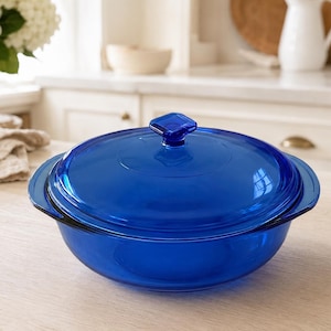 May include: A round, deep, cobalt blue glass casserole dish with a matching transparent lid. The lid features a square-shaped knob handle at the center, and the base has two integrated side handles for lifting. The glass is smooth and translucent.