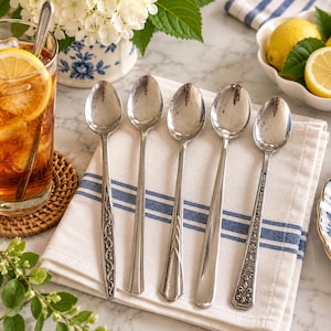 May include: A set of five silver-toned metal spoons arranged in a row on a white cloth napkin with blue stripes. Each spoon features a distinct handle design, including floral, geometric, and scrollwork patterns, with polished oval bowls.