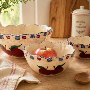 May include: Three off-white ceramic bowls with scalloped edges, decorated with red apples and blue accents. The bowls vary in size, with the largest bowl measuring approximately 12 inches in diameter. One bowl contains a red apple.