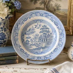 Peut inclure: Une assiette ronde en céramique ornée d'un motif bleu et blanc de type saule. Le centre représente un paysage avec une pagode, un pont et des oiseaux, tandis que le bord est décoré d'une bordure géométrique. L'assiette est sur un support.