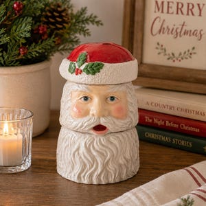 May include: A ceramic Santa Claus figurine featuring a red hat with a white brim, green holly leaves, and red berries. The figure has a detailed face with blue eyes, rosy cheeks, and a textured white beard with an open mouth at the center.
