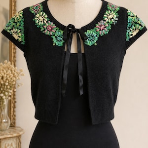 May include: A black short-sleeved bolero cardigan featuring a round neckline with a black ribbon tie closure. The garment is decorated with embroidered floral patterns in shades of green, pink, and cream, accented with small shimmering sequins.