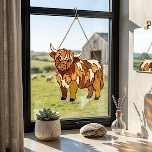 May include: A decorative stained glass Highland cow, hanging in a window. The cow is in shades of brown, tan, and white, with a chain for hanging. The background shows a rural landscape with a barn and sheep grazing in a field.