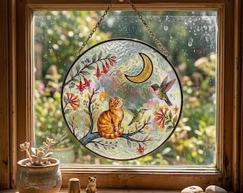 Op de afbeelding: Een ronde glas-in-lood raamdecoratie met een kat, kolibries en een maan. De kat is oranje en wit, zittend op een tak met rode en oranje bloemen. Twee kolibries zijn afgebeeld, een groene en een met rode accenten. Het raam wordt opgehangen door een gouden ketting.