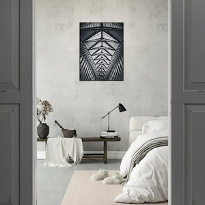 May include: A black and white architectural photograph of a building's interior, featuring a repeating geometric pattern of beams and trusses. The image is framed on a light gray wall above a bench with a blanket, a lamp, and a bed with white bedding.