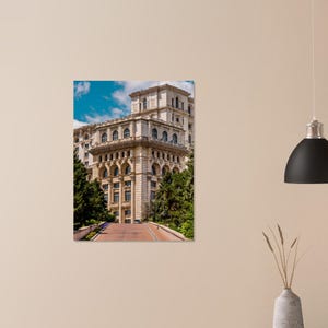 May include: A large, multi-story building with a beige facade and numerous windows. The building has a classical architectural style, with arched windows and decorative elements. A red pathway leads to the building, surrounded by green trees and a blue sky.