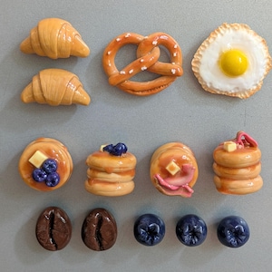 May include: A collection of food-themed magnets including two golden croissants, a brown pretzel with salt, a fried egg, four stacks of pancakes with butter and toppings, two dark brown coffee beans, and three round blue blueberries.