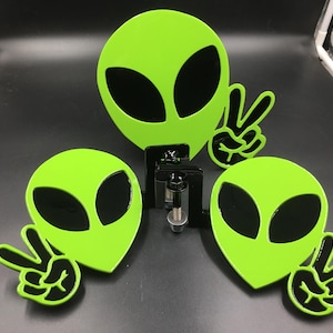 May include: Three neon green alien head decorations with black eyes and a peace sign hand gesture. The alien heads are mounted on a black metal bracket. The background is black.