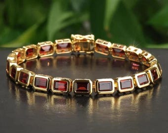 May include: A gold bracelet featuring rectangular, deep red gemstones. The bracelet is composed of individual links, each holding a gemstone. The clasp is also gold. The bracelet is displayed on a dark surface with a blurred green background.