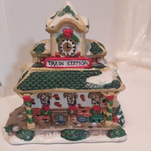 Puede incluir: Figura de cerámica de estación de tren navideña con techo verde, detalles de nieve y una torre de reloj. El letrero dice "TRAIN STATION". El edificio tiene paredes de ladrillo rojo y detalles decorativos, creando una escena festiva.